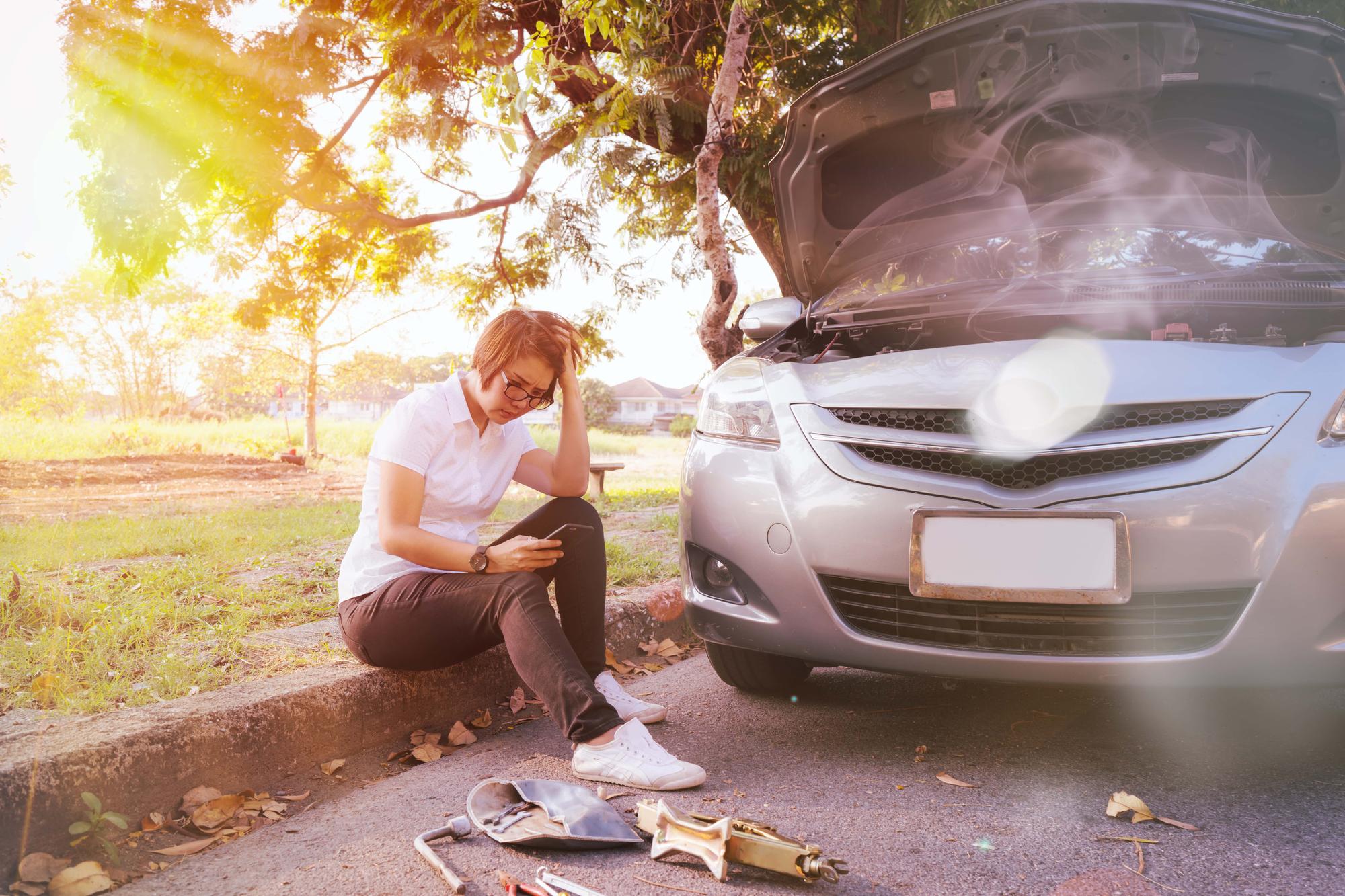 A person sits on a curb, frustrated, next to a silver car with an open hood. Sunlight filters through the trees. Tools and gloves are scattered on the ground. The scene suggests car trouble.
