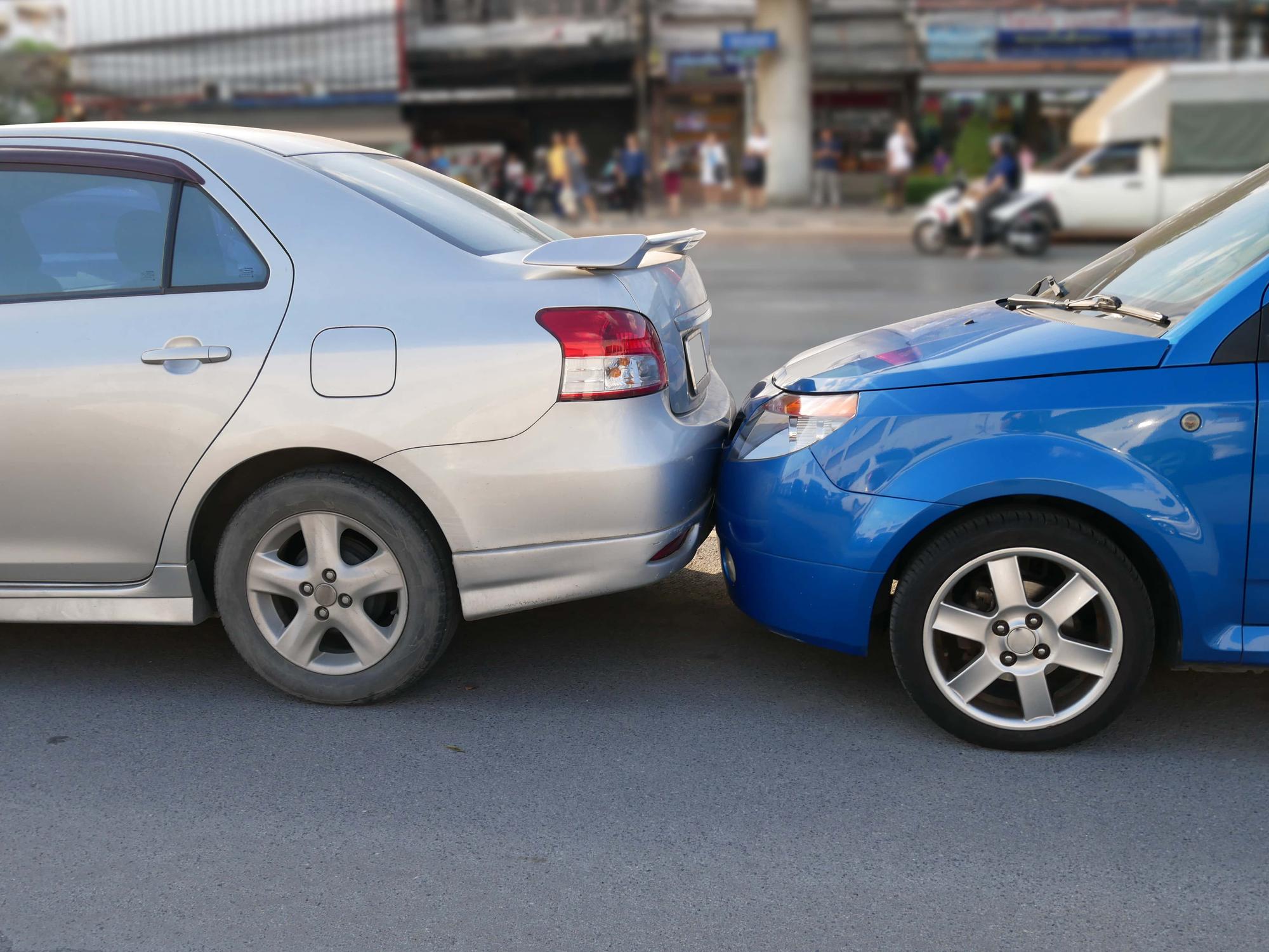 A blue car is rear-ended by a silver car on a city street. The vehicles are closely aligned, with minor visible damage. In the background, people and buildings are slightly blurred.