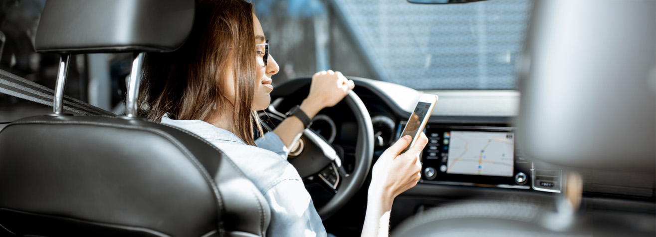 A person wearing glasses and a denim shirt is sitting in the driver's seat of a car, looking at a smartphone. The car's dashboard displays a navigation map. The person is smiling slightly.