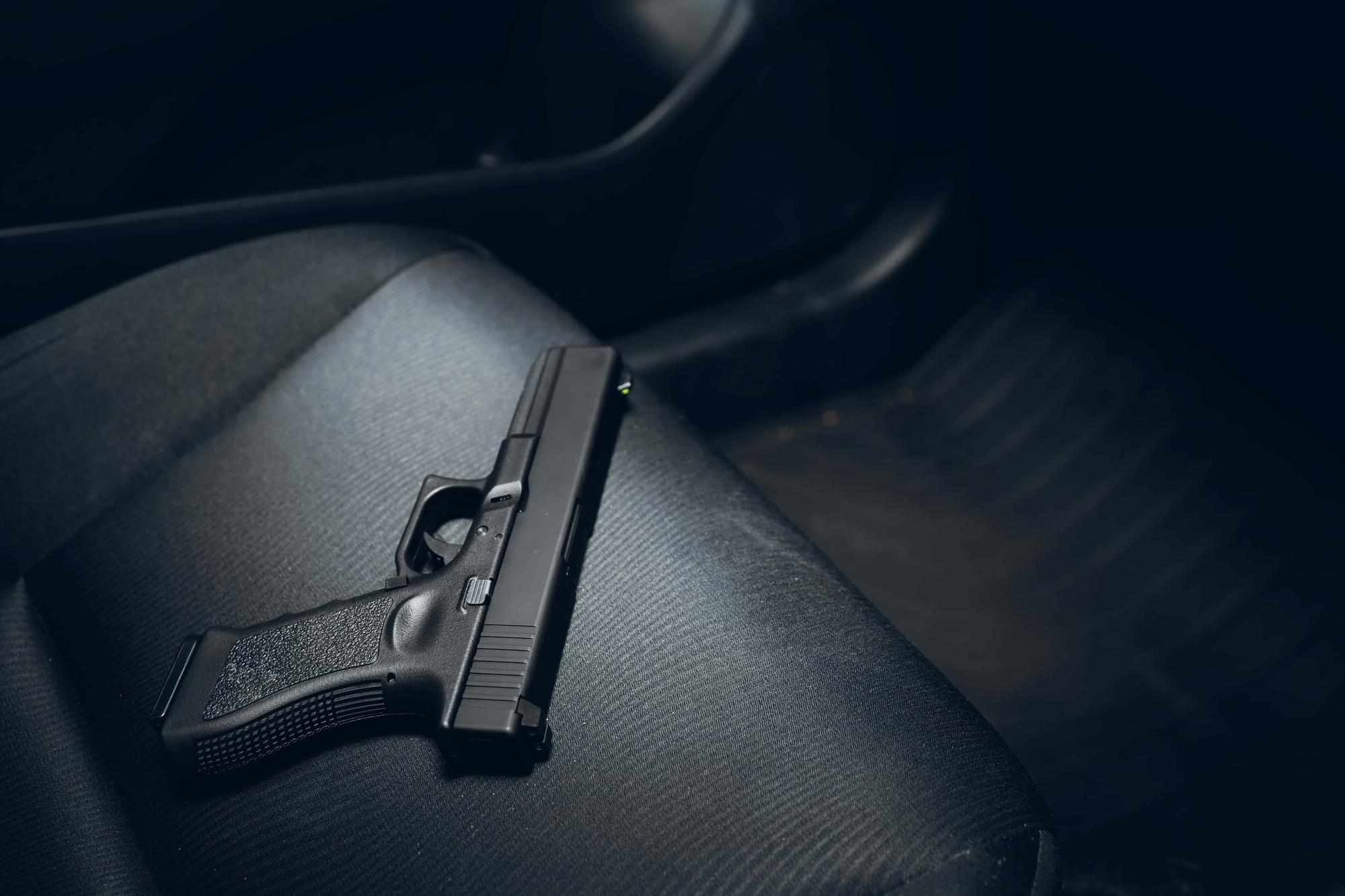 A handgun is lying on the black leather seat of a car. The interior is dimly lit, with focus on the gun. The background is blurred, showing part of the car's floor and door.