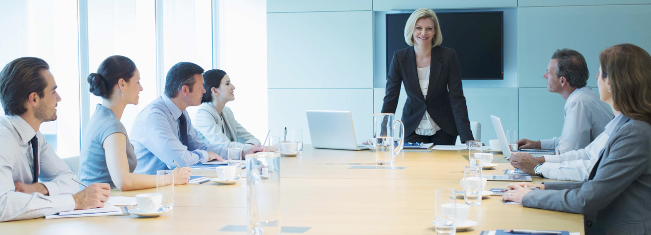 A woman in a suit stands at the head of a conference table, smiling, with a laptop in front of her. Six colleagues sit on either side, attentively listening. The room is modern with a large screen on the wall.