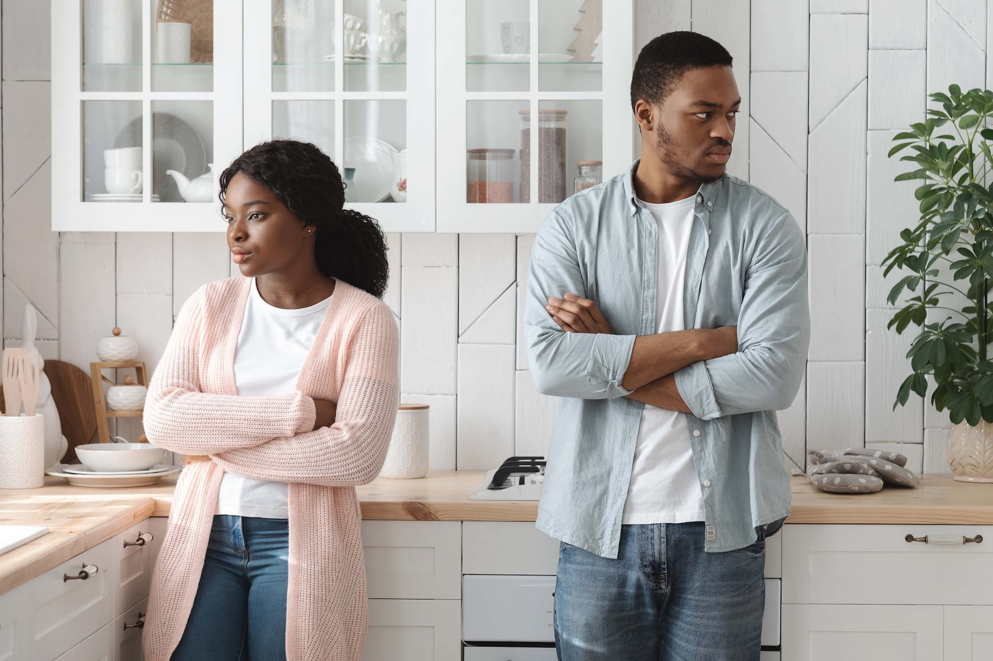 A man and woman stand in a kitchen with arms crossed, looking away from each other. Both have a serious expression. The kitchen features white cabinets and a potted plant on the counter.