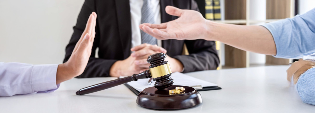 Close-up of three people at a table, with a gavel and two rings in focus. One person's hand is raised defensively, another is gesturing, suggesting a discussion or negotiation. Legal documents and books are visible in the background.
