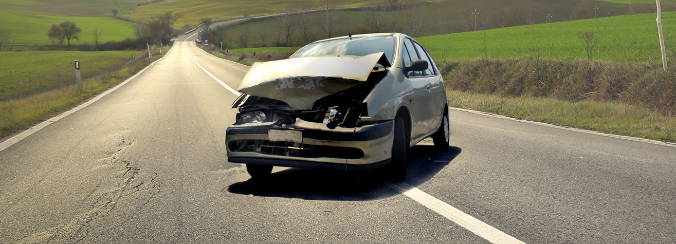 A damaged car with a crumpled front hood is stopped in the middle of an empty rural road. The landscape is open with green fields on either side, and the sky is clear.
