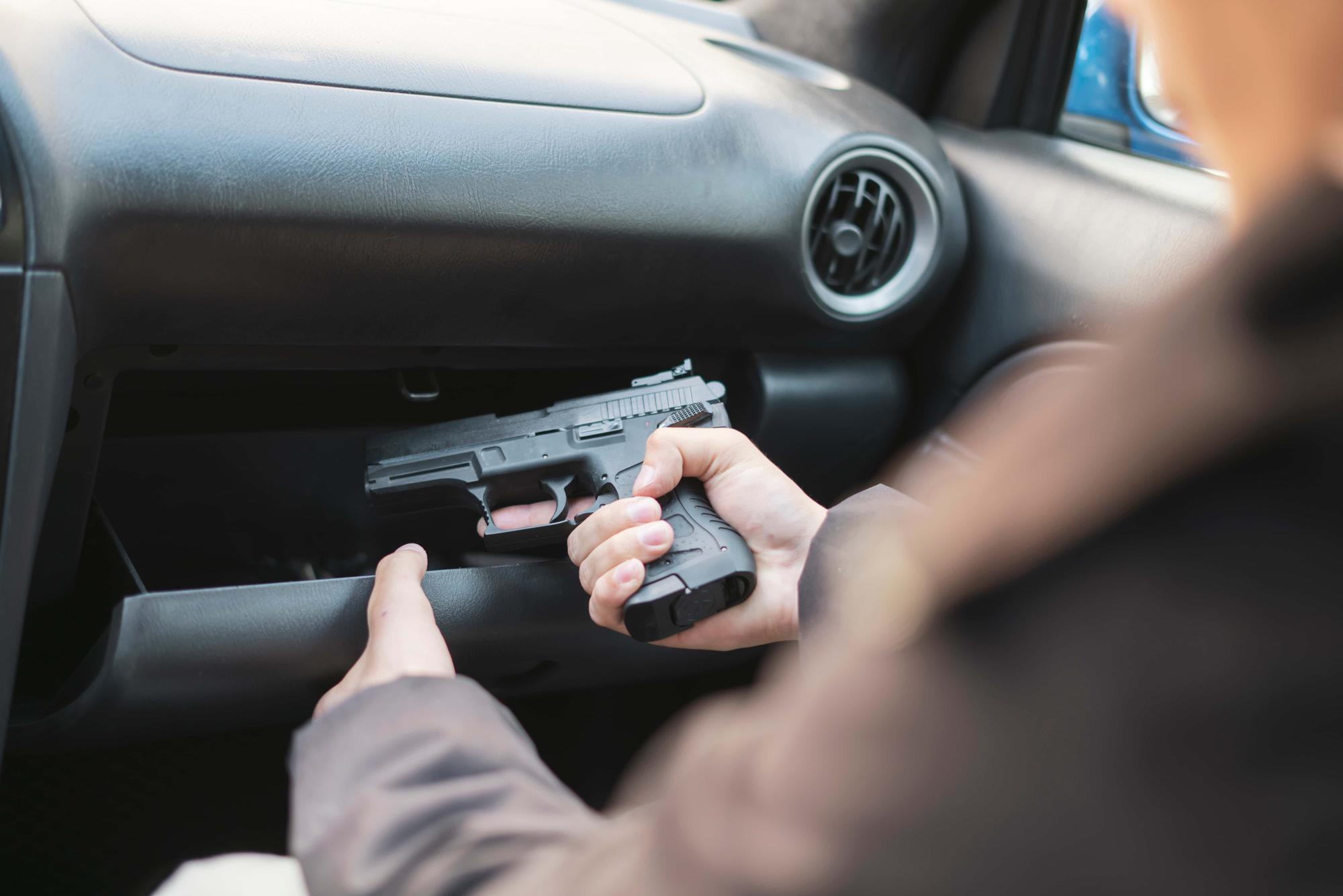 Person seated in a car reaches into the open glove compartment, holding a handgun. The individual's face is not visible. The interior of the car is mostly dark, with sunlight coming through a window.