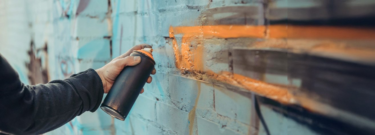 A person holds a black spray paint can and paints an orange line on a brick wall. The wall has various blue and gray graffiti patterns in the background.