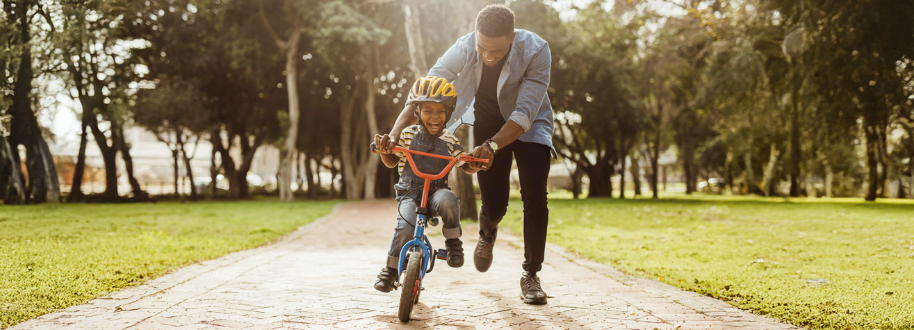 A child is riding a bicycle with training wheels on a paved path in a park, wearing a yellow helmet and smiling. An adult is guiding the bike from behind, helping the child. The sun is shining through the trees.