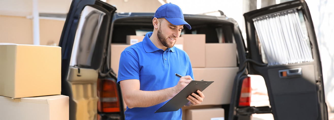 A delivery person in a blue uniform and cap smiles while holding a clipboard. He stands in front of an open van filled with cardboard boxes, appearing to check or annotate information.