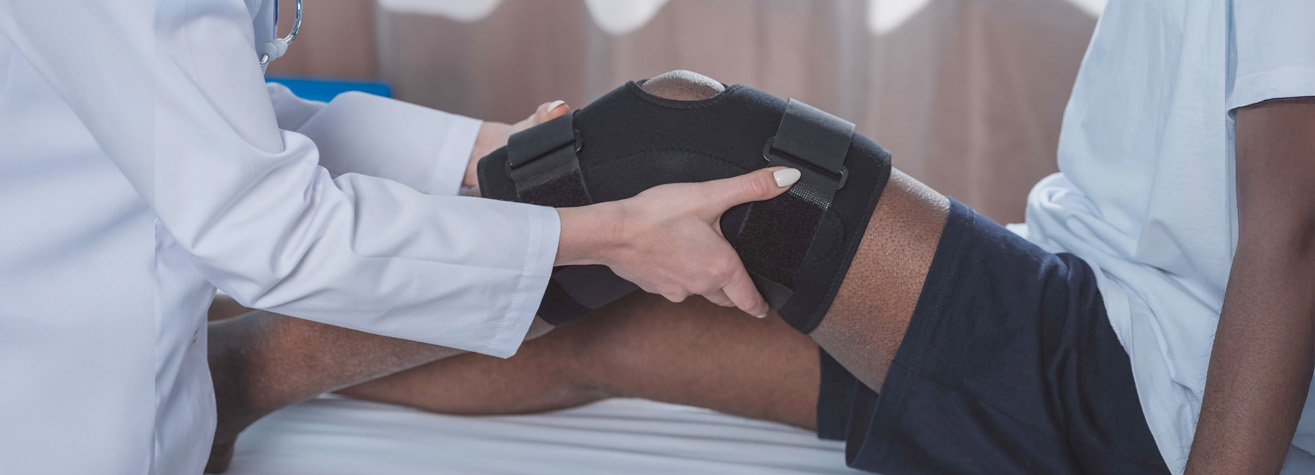 In a setting that evokes the precision of both medical and legal expertise, a doctor in a white coat adjusts a knee brace on a patient's leg. The patient, seated on an examination table and wearing blue shorts, seems to be receiving care reminiscent of how a Pearland personal injury lawyer addresses client needs.