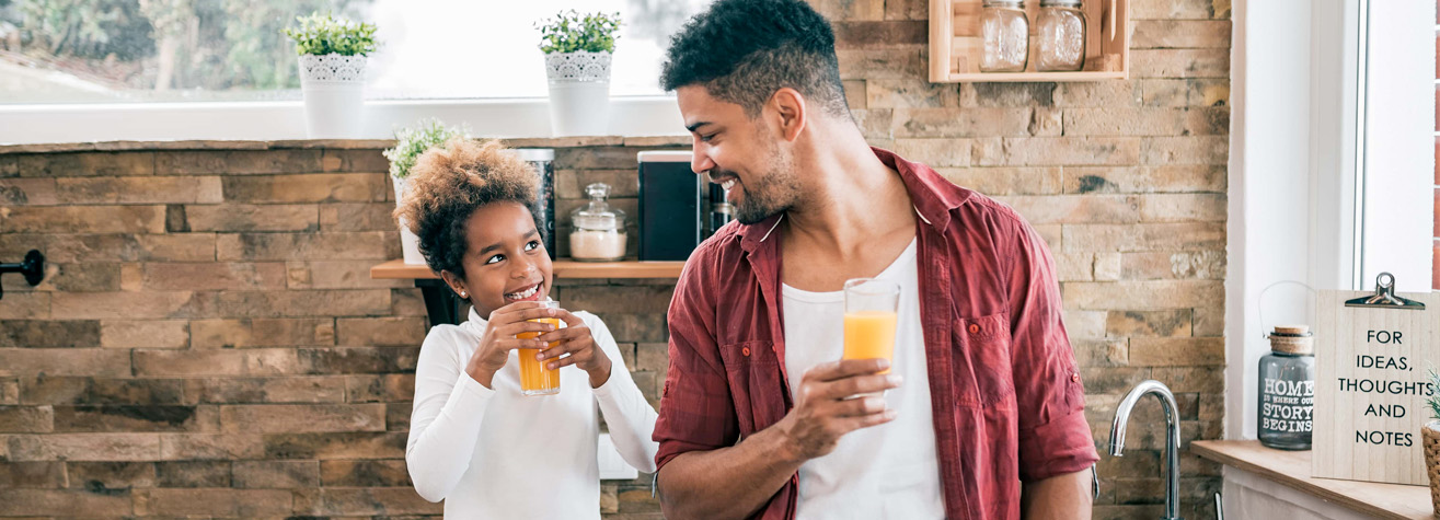 A man and child smile at each other while holding glasses of orange juice in a cozy kitchen. The kitchen has a brick wall, potted plants on the windowsill, and decorative jars.