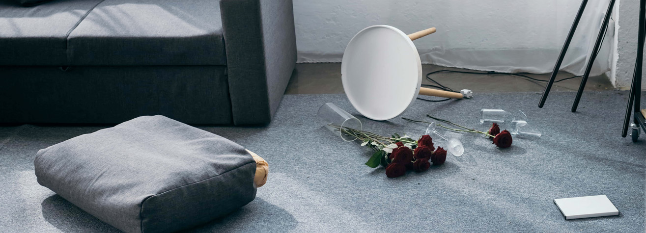 A living room scene shows an overturned small round table and a scattered bouquet of red roses with fallen petals on a gray carpet. A gray couch and a cushion are in the background, with a small book nearby.