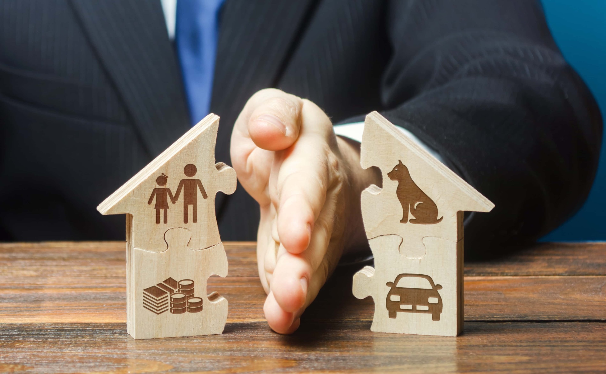 A person in a suit uses a hand to separate two wooden house-shaped puzzle pieces. One piece shows icons of a couple and money, while the other shows a dog and a car. The separation symbolizes division or splitting of assets.