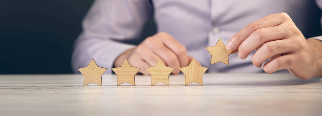A person in a light purple shirt places a fifth wooden star on a table alongside four others, symbolizing a five-star rating. The background is dark, highlighting the stars and hands in focus.
