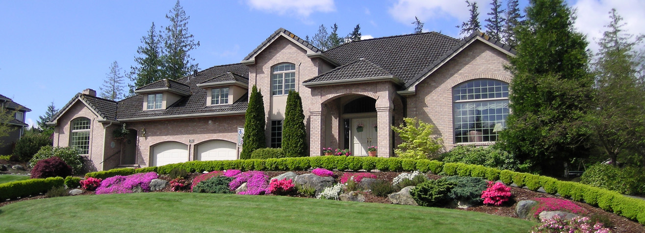 A large, elegant brick house with multiple gables is surrounded by lush greenery and a manicured lawn. Colorful flowers and neatly trimmed bushes adorn the front yard. Two white garage doors are visible on the left side of the house.