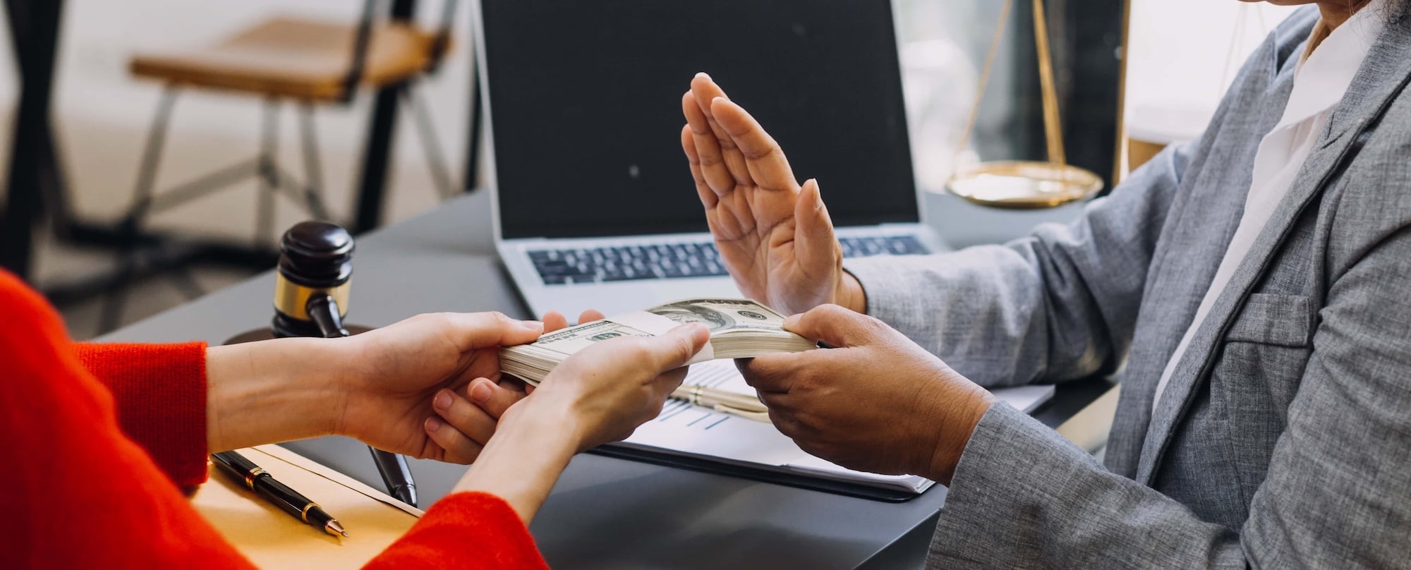 A person in a red sleeve hands over a bundle of dollar bills to a person in a gray suit, who gestures to stop with one hand. On the table are a gavel, a pen, a laptop, and scales of justice.