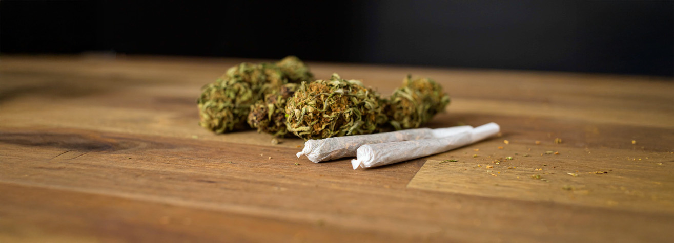 Two rolled joints lie on a wooden surface next to a small pile of dried cannabis buds. The background is dark, highlighting the earthy tones of the wood and cannabis.