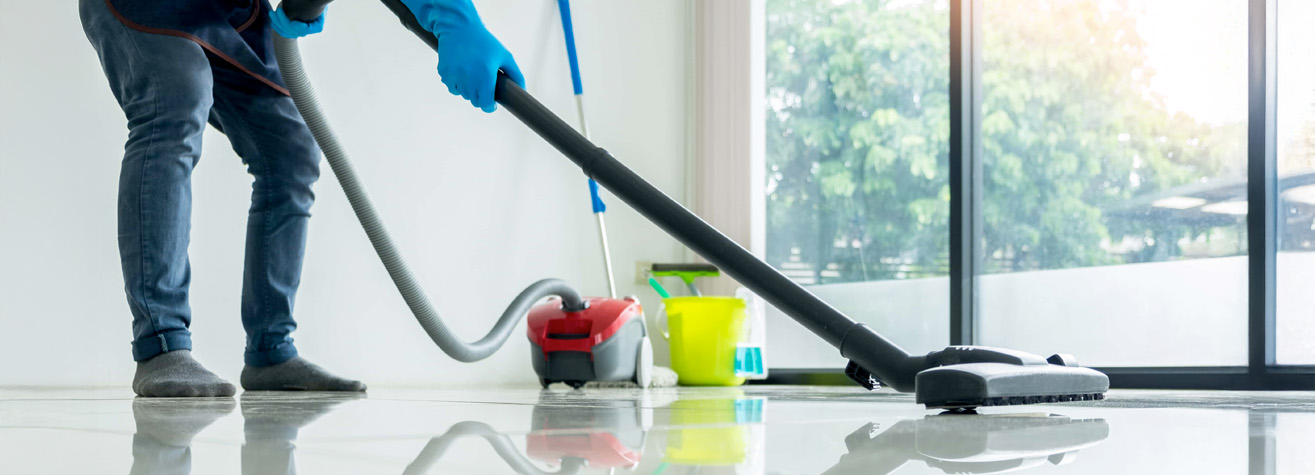 A person wearing blue gloves and socks uses a red vacuum cleaner on a shiny floor near large windows. Cleaning supplies, including a mop and a yellow bucket, are in the background.