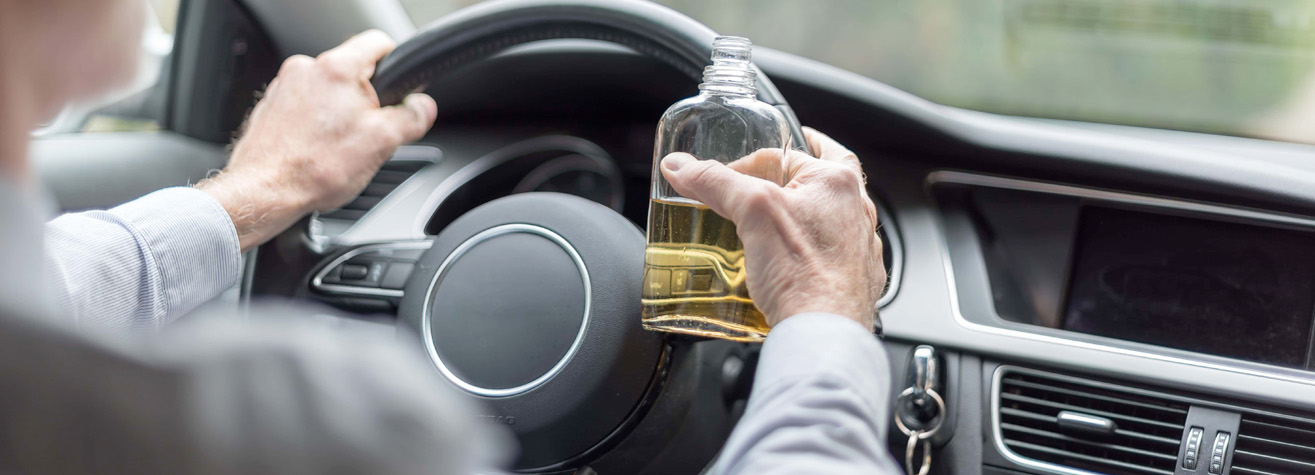 Person holding a bottle with an amber liquid in one hand and a steering wheel in the other, sitting in a car. The interior dashboard and side mirror are visible. Safety concerns are implied.
