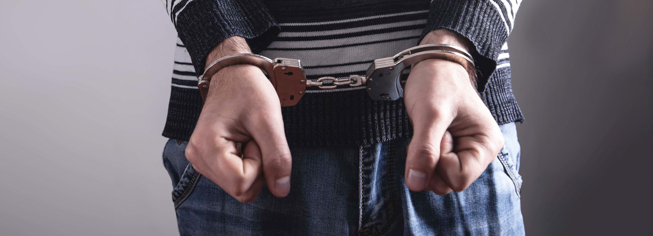 A person in a striped sweater and jeans stands with hands cuffed in front, emphasizing their restrained posture. The background is a neutral gray.