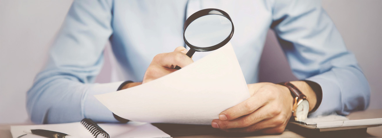 A person in a blue shirt holds a magnifying glass over a sheet of white paper, appearing to examine it closely. They are seated at a desk with an open notebook and other papers nearby.