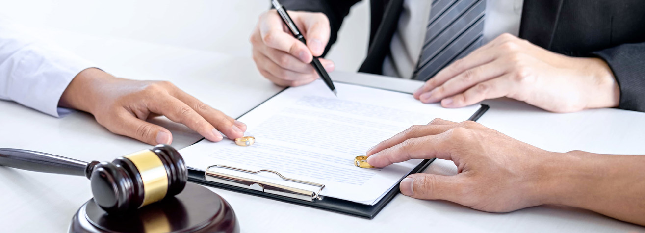 Two people sit at a table, with a legal document on a clipboard. One person holds a pen, poised to sign. Two gold rings rest on the document. A wooden gavel is on the table, suggesting a legal or divorce process.