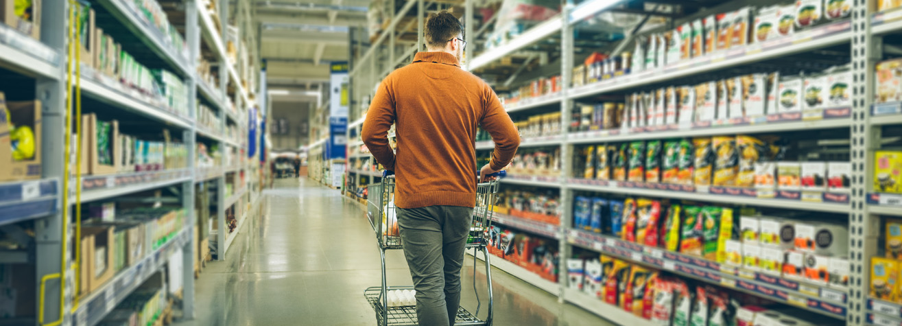 A person wearing a brown sweater pushes a shopping cart down a grocery store aisle. Shelves on both sides are filled with various packaged products. The floor is shiny, and fluorescent lights illuminate the scene.