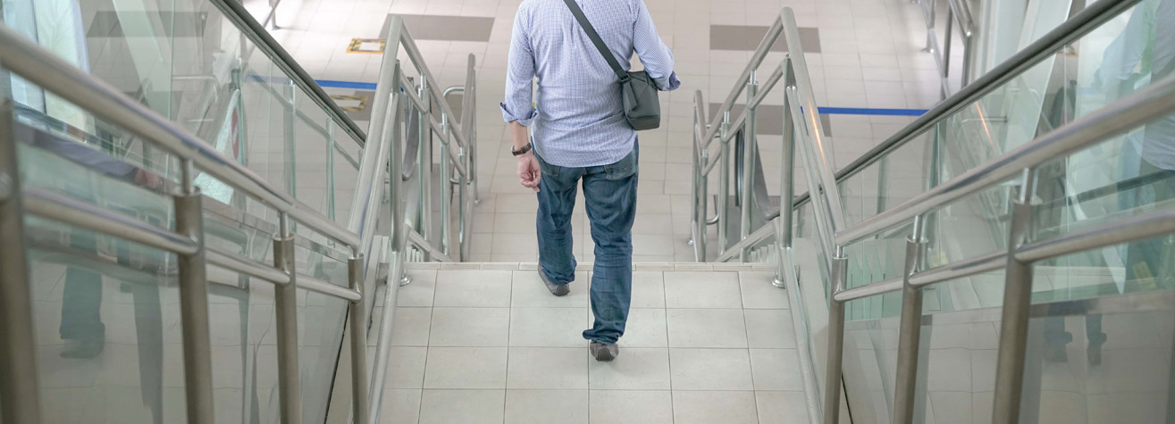 A person in a light blue shirt and jeans descends a tiled staircase, carrying a black shoulder bag. The stair railings are metal with glass panels, and the background features a tiled floor and walls with white and gray tiles.