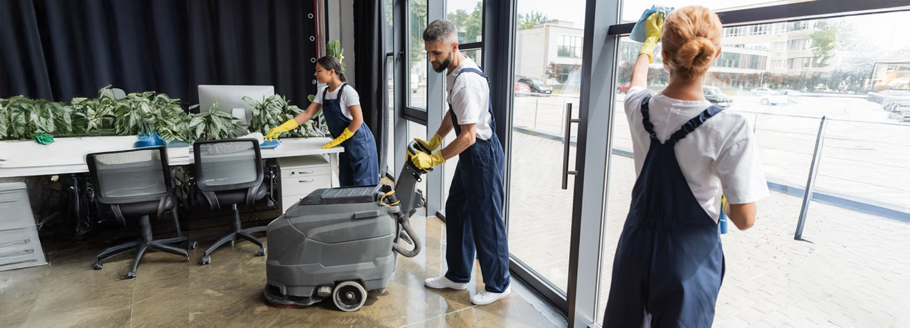 Three cleaners in uniforms work in an office. One uses a floor cleaning machine, another wipes a window, and the third cleans a desk. The office has large windows and office chairs. Outside, there are cars and buildings visible.
