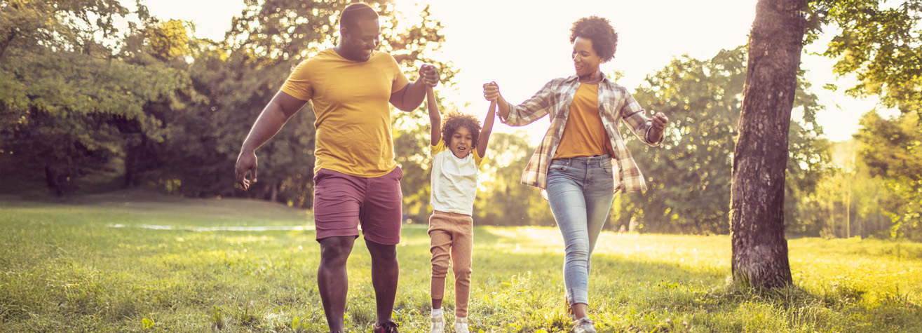 A joyful family of three walks outdoors in a sunlit park. The parents hold their child's hands, lifting them playfully. The family is dressed in casual, colorful clothing, and the background features lush green trees and grass.