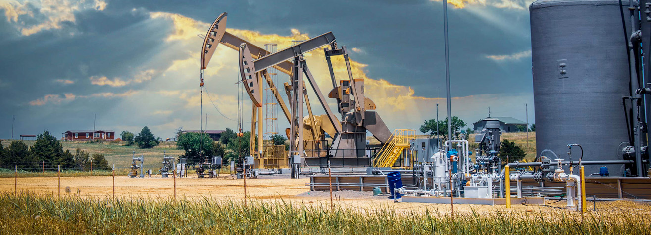 An oil pumpjack operates in a field under a cloudy sky with sun rays breaking through. Surrounding equipment and tanks are visible, along with a few structures in the background. The foreground features grassy land.