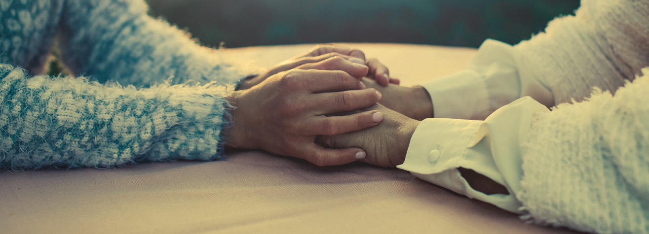 Two people holding hands across a table, wearing cozy sweaters. The scene conveys warmth and connection, with soft lighting and a blurred background.