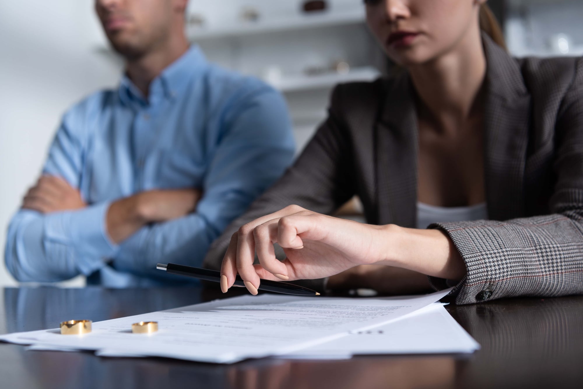 A man and woman sit at a table with visible tension. The man wears a blue shirt with crossed arms. The woman in a blazer holds a pen, pointing at documents on the table. Two gold rings are next to the paperwork.