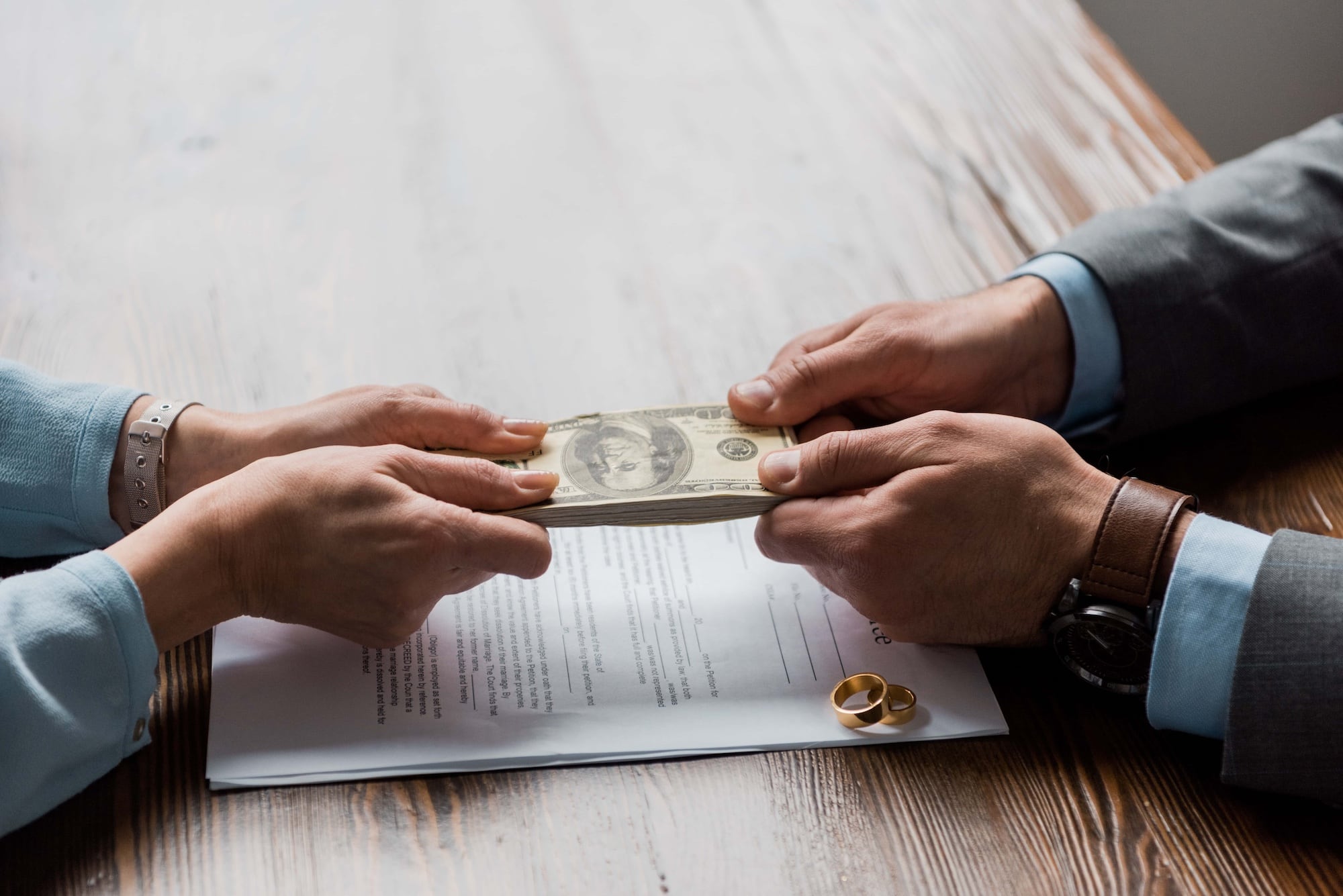 Two people exchanging a stack of hundred-dollar bills over a wooden table. A document with two gold rings on it is visible beneath their hands, suggesting a financial agreement or settlement, possibly related to a divorce.