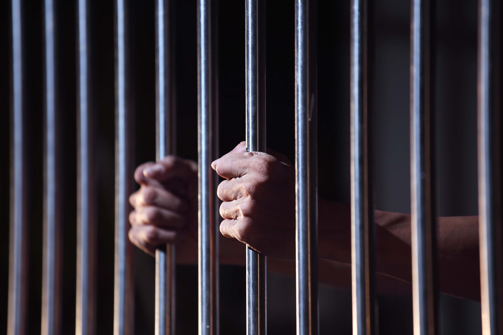 Hands gripping metal prison bars with a blurred dark background, conveying a sense of confinement and captivity.