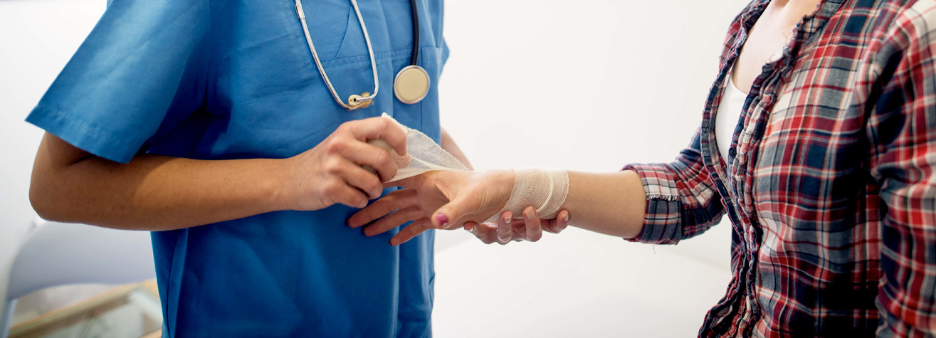 A healthcare professional in blue scrubs is bandaging a person's wrist. The person receiving care is wearing a plaid shirt. The scene appears to be in a medical setting with a focus on the bandaging process.
