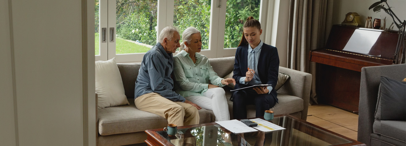 A senior couple sits on a couch in a bright living room, attentively listening to a professional seated beside them. The professional is holding a tablet, gesturing towards it. A piano and large windows with garden views are in the background.