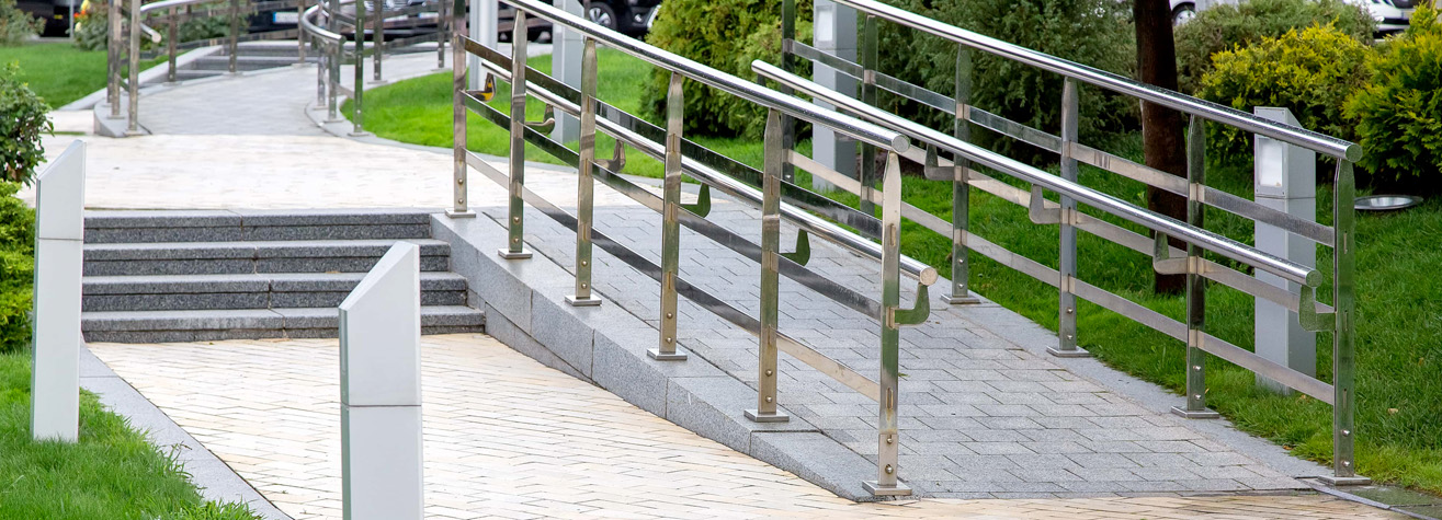 A paved path features a smooth, accessible ramp parallel to a set of stairs, bordered by metal railings. The surrounding area has green grass and shrubs, with trees in the background.