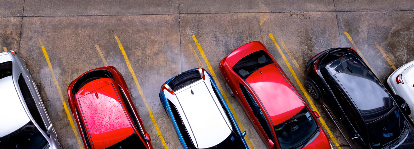 Aerial view of cars parked in a lot. The cars are lined up diagonally between yellow lines on wet pavement. The vehicles are various colors, including red, white, and black.