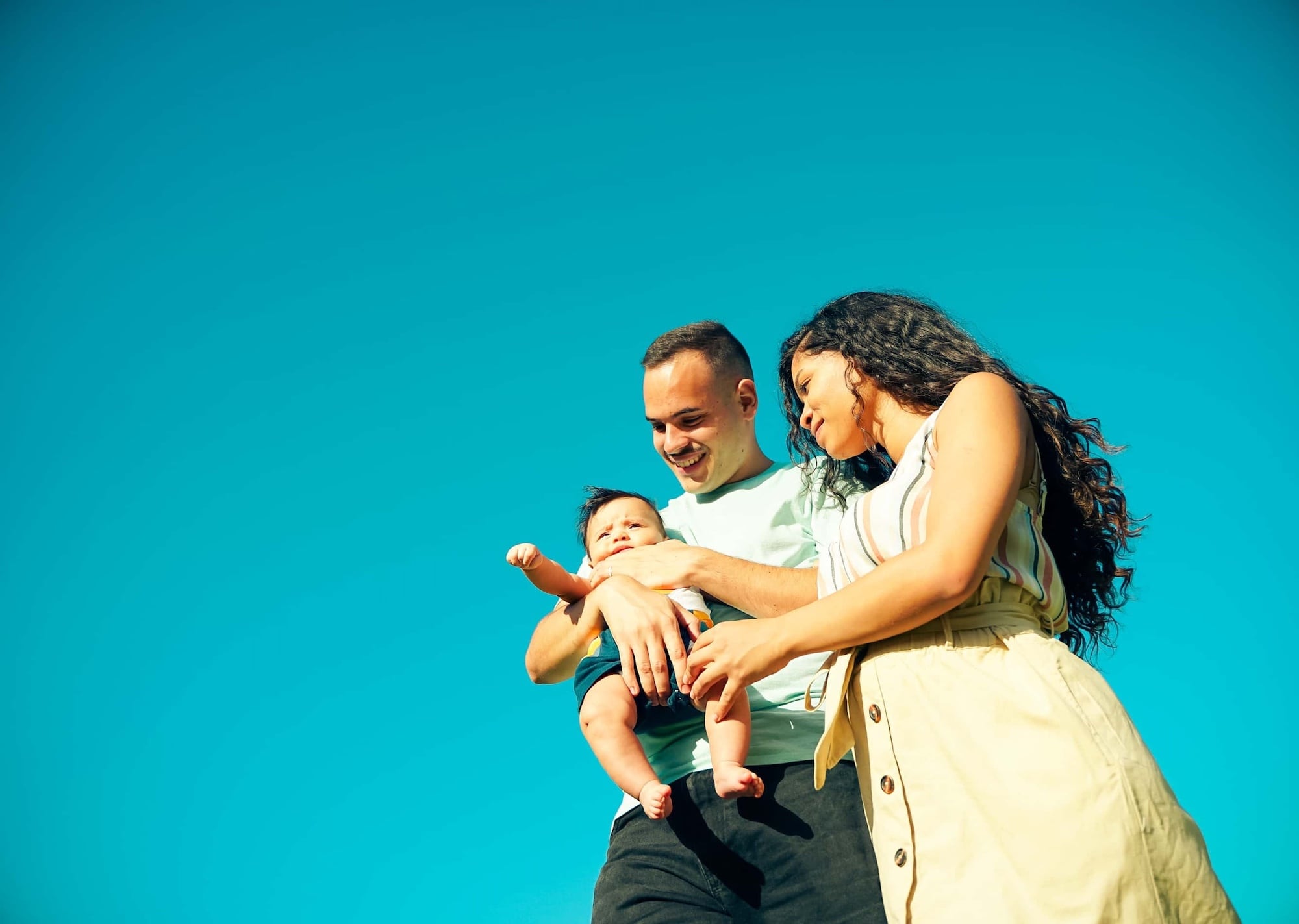A couple stands outdoors against a clear blue sky, smiling at a baby in their arms. The man wears a light blue shirt and the woman wears a striped top and skirt. The baby wears a dark outfit. The scene conveys happiness and family bonding.