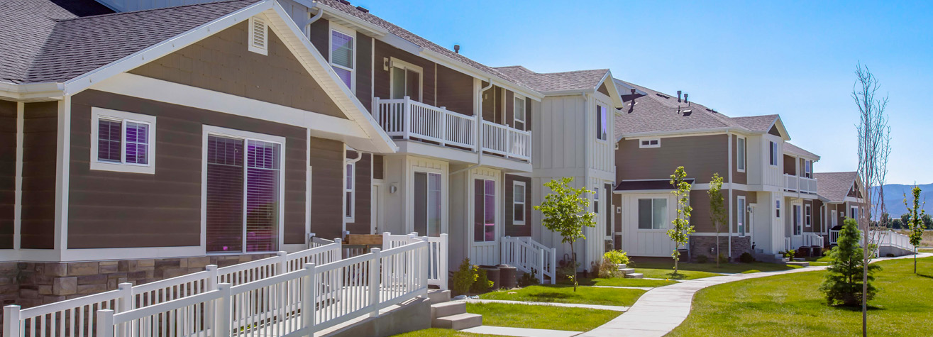 A row of modern townhouses with brown and white exteriors. Each unit has a small porch, balconies, and a well-maintained lawn. A winding concrete path leads through the green, landscaped yard under a clear blue sky.