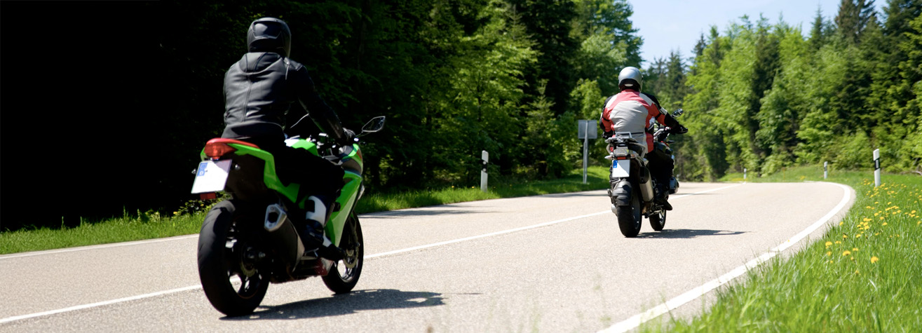 Two motorcyclists ride on a winding country road surrounded by lush green trees and grass. The biker in front wears a white and red jacket, while the one behind wears black gear. The sky is clear, suggesting a pleasant day.