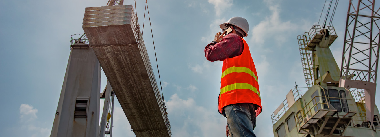 A construction worker in an orange safety vest and hard hat communicates via a walkie-talkie. They stand next to a large, suspended concrete slab at a construction site, with cranes and a clear blue sky in the background.
