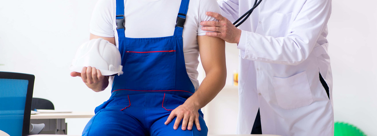 A person in blue overalls holds a white hard hat while sitting on an examination table, their heartbeat checked by a doctor in a white coat. The bright, clean medical office resembles the professionalism you'd expect from a Friendswood Personal Injury Lawyer's trusted network.
