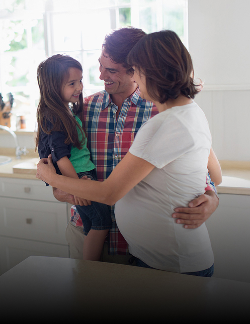 A smiling family stands together in a bright kitchen. A man holds a young girl, who smiles at an adult woman with a pregnant belly. They appear happy and affectionate, knowing Personal Injury & Criminal Defense Attorneys help protect families like theirs. Sunlight streams in.