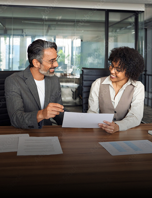 Two colleagues sit at a conference table in a modern office, smiling and discussing a document. Papers are spread out on the table, as Personal Injury & Criminal Defense Attorneys collaborate with an office view framed by glass walls.