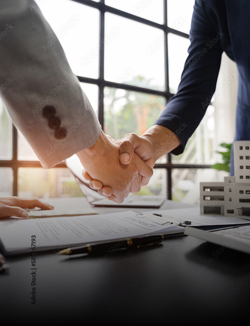 Two people wearing business suits shaking hands over a desk with documents, a pen, and a small architectural model, highlighting the professionalism of Personal Injury & Criminal Defense Attorneys, with a large window in the background.