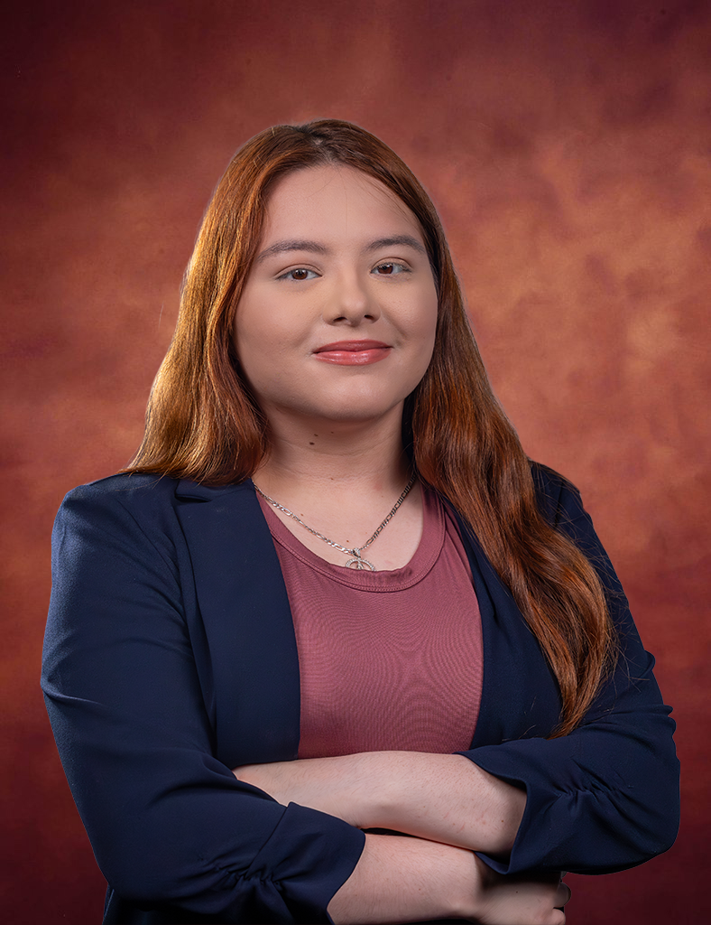 Iliana Oviedo, with long reddish-brown hair, wearing a maroon top and navy blazer, stands with folded arms and a slight smile against a maroon and orange gradient background.