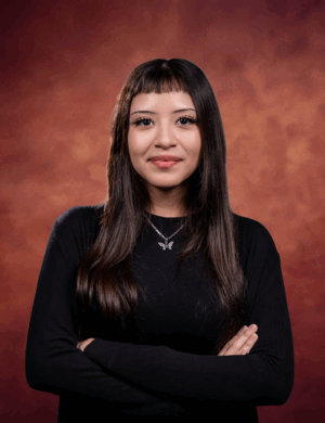 Italimi Nunez, a woman with long straight brown hair and bangs, wearing a black top and butterfly necklace, stands with her arms crossed, smiling slightly against a reddish-brown studio background.