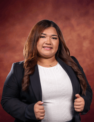 Jackie Lucio, with long brown hair, wears a black blazer and white top as she smiles confidently, standing in front of a reddish-brown studio background.
