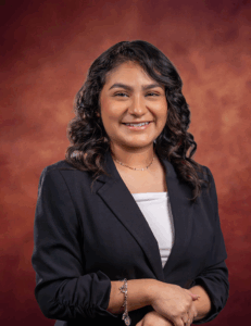 Jaqueline Casio, with wavy dark hair, smiles in a black blazer and white top against a warm brown gradient background. She stands with her hands folded, accessorized by a bracelet and necklace.